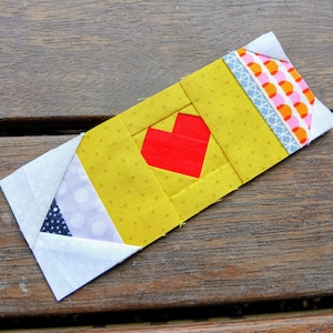 May include: A handmade fabric pencil-shaped item with a red heart in the center. The pencil is yellow with a white tip, a blue and white band, and a patterned eraser end. The item is placed on a wooden surface.