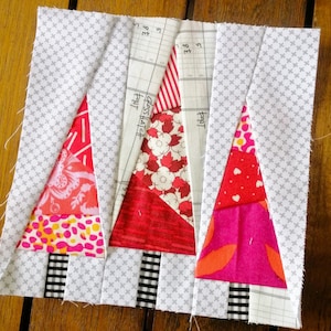 May include: Three red and pink patchwork fabric Christmas trees with black and white checked fabric bases on a white background with a grey grid pattern. The fabric trees are made from different patterns of red, pink, and white fabric.