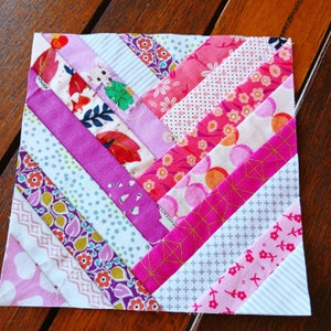 May include: A pink, purple, and white patchwork quilt block with a chevron pattern. The block is made up of strips of fabric sewn together in a diagonal pattern. The fabric has a variety of patterns, including floral, geometric, and polka dots.