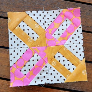 Op de afbeelding: Een vierkant patchwork quiltblok met een geometrisch ontwerp. Het blok heeft secties van witte stof met bruine stippen, roze stof met grote oranje cirkels en gouden stof met kleine witte patronen. Het blok is gemaakt van aan elkaar genaaide stukken stof.