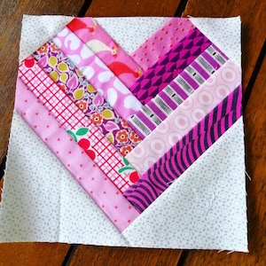 May include: A pink and purple patchwork heart quilt block with a white background. The block is made up of strips of fabric sewn together in a diagonal pattern.