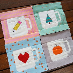 May include: Four colorful fabric squares with a mug design. Each mug features a different design: a pencil, a Christmas tree, a red heart, and a pumpkin. The squares are framed with patterned fabric in pink, green, blue, and gray.