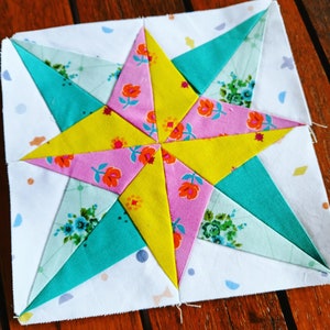 May include: A patchwork quilt block featuring a star design with yellow, pink, and teal fabric. The star is surrounded by white fabric with a subtle pattern.