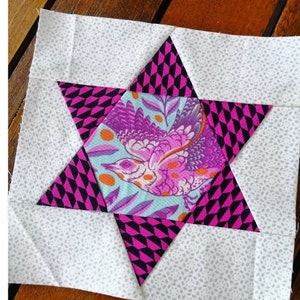 Pode incluir: Um quadrado de tecido branco com um design de estrela rosa e preta. A estrela tem uma peça central de tecido com um design de pássaro roxo e laranja sobre um fundo azul.