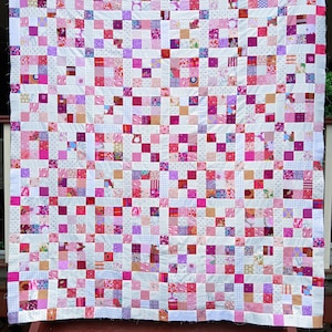 May include: A handmade quilt featuring a patchwork design with a variety of pink, purple, and white fabrics. The quilt is composed of numerous small squares, creating a visually appealing and intricate pattern. The quilt is displayed outdoors.