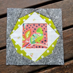 May include: A square fabric block with a bird design. The central square features a green bird on a branch against a pink background. It is surrounded by white, green polka dot, and black and white patterned fabrics. The block is likely for quilting.
