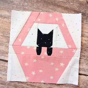 May include: A square fabric quilt block featuring a pieced design of a black cat sitting inside a pink basket. The basket is constructed from pink fabric with a white star pattern, set against a white background with small colourful dots.