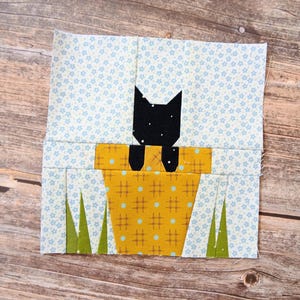 Peut inclure: Un bloc de courtepointe en tissu carré représentant la silhouette d'un chat noir sortant d'un pot de fleurs jaune moutarde à motif de grille marron et points bleus. Le fond est bleu clair avec de petites fleurs blanches et des brins d'herbe verts.