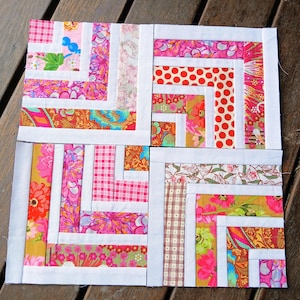May include: Four colorful quilt blocks with a log cabin design. Each block features a variety of patterned fabrics in pink, red, green, and brown, framed by white borders. The blocks are arranged on a wooden surface.