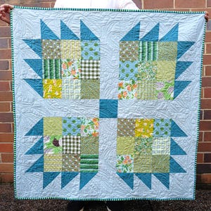 May include: A handmade quilt with a light blue background and a patchwork design. The quilt features teal triangles and squares of various green and floral patterns. The border is light blue with a green and white striped trim. The quilt is approximately 102 cm square.