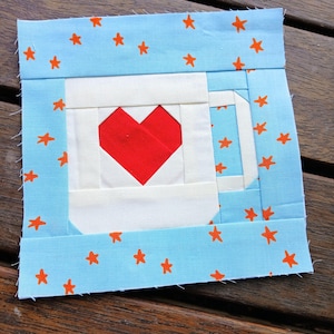 May include: A fabric quilt block featuring a white mug with a red heart in the center. The mug is set against a light blue background adorned with orange star shapes. The block is a square shape.