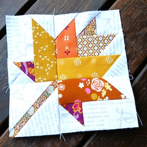 May include: A colorful patchwork quilt block featuring a starburst design. The block incorporates various fabrics in shades of orange, yellow, and purple, with floral and geometric patterns. The background is white with printed text.