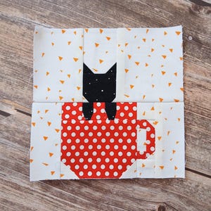 Peut inclure: Un bloc de courtepointe en tissu carré représentant une tasse à café rouge à pois blancs avec une anse sur le côté droit. Une silhouette de chat noir aux oreilles pointues est placée dans la tasse. Le fond est en tissu blanc à petits triangles orange.