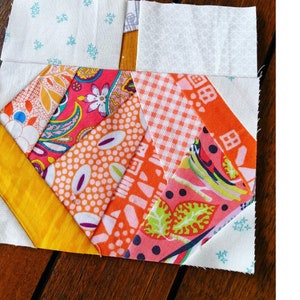 Può includere: Un blocco di trapunta patchwork con una forma di cuore al centro. Il cuore è fatto di diversi tessuti in tonalità di arancione, rosa e bianco con motivi diversi. Lo sfondo è un tessuto bianco con un piccolo motivo a stelle blu.