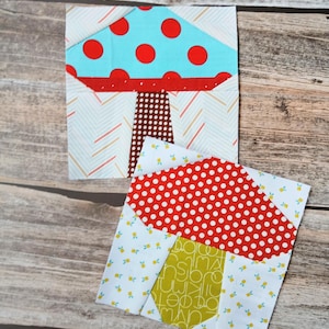 Pode incluir: Dois blocos de tecido quadrados para colcha com desenhos de cogumelos. O bloco superior tem um chapéu azul-claro com bolinhas vermelhas e um caule castanho com bolinhas. O bloco inferior tem um chapéu vermelho com bolinhas brancas e um caule verde com letras brancas.