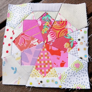 Op de afbeelding: Een patchwork quiltblok met een stervormig ontwerp. Het blok is gemaakt van verschillende stoffen in tinten roze, rood, wit en geel. De stoffen hebben verschillende patronen, waaronder stippen, bloemenprints en geometrische ontwerpen.