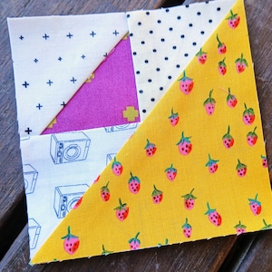 Peut inclure: Un bloc de patchwork carré en tissu avec quatre sections. Une section a un fond jaune avec des fraises roses. Une autre a un fond crème avec des signes plus noirs. La troisième a un fond crème avec des pois noirs. La quatrième a un fond blanc avec des illustrations de caméras bleues.