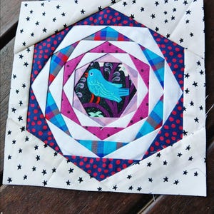 May include: A quilt block featuring a blue bird in the centre surrounded by concentric rings of pink, white, blue and red fabric. The outer border is white with black stars.