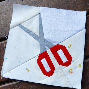 May include: A white fabric square with a red and white geometric design. The design is made up of two red shapes with white stripes, and a gray and white pattern in the background.