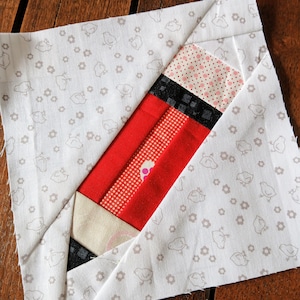 May include: A red, white, and black fabric block with a geometric pattern. The block is made of several different fabrics sewn together. The block is on a white background with a pattern of small gray birds.