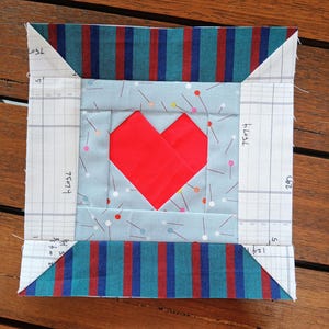 Könnte beinhalten: Ein handgefertigter Quiltblock mit einem roten Herzdesign. Das Herz ist auf einem hellblauen Stoff mit winzigen Nadelgrafiken zentriert. Der Block ist mit gestreiftem Stoff in Türkis, Rot und Weiß umrahmt und enthält weiße Ecken mit handschriftlichem Text.