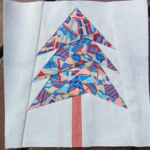 May include: A patchwork quilt block featuring a stylized Christmas tree with a red, blue, and pink geometric pattern. The tree is on a white background with a small, repeating pattern.