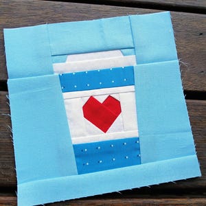 May include: A square fabric block featuring a coffee cup design with a red heart. The cup has a white lid, blue polka dot pattern, and a red heart. The block is framed in light blue fabric, perfect for quilting projects.