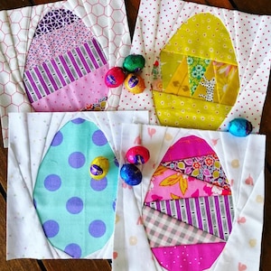 Può includere: Quattro blocchi di patchwork a forma di uovo di Pasqua fatti a mano con uno sfondo bianco e disegni di patchwork colorati. Ogni blocco presenta un motivo e uno schema di colori diverso, tra cui rosa, viola, giallo, verde, blu e bianco. I blocchi sono circondati da piccoli ovetti di Pasqua di cioccolato.