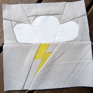 May include: A gray and white fabric quilt block with a yellow lightning bolt and a white cloud. The cloud is made of fabric and is layered on top of the gray fabric.