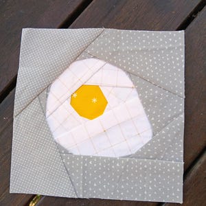 May include: A square fabric quilt block with a fried egg design. The yellow yolk is a hexagon, surrounded by white fabric. The block is framed by gray patterned fabric. The design is suitable for quilting projects.