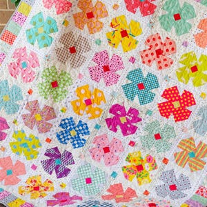 May include: A handmade quilt featuring a repeating floral pattern. Each flower is composed of colorful fabric patches in various patterns, including polka dots, gingham, and stripes. The quilt has a white background and a scalloped edge.