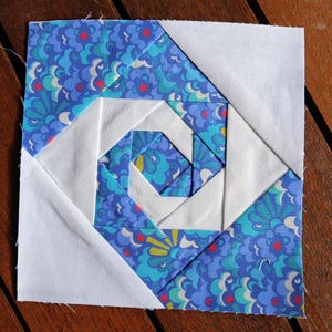 Könnte beinhalten: Quadratischer Stoff-Quiltblock mit einem zentralen Spiraldesign. Der Block kombiniert weißen und gemusterten blauen Stoff mit floralen und Blattmotiven. Das Design ist geometrisch, mit präzisen Winkeln und Nähten.