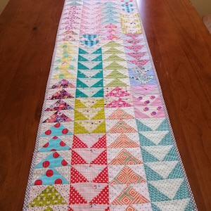 K&ouml;nnte beinhalten: Ein farbenfroher Patchwork-Tischl&auml;ufer mit einem geometrischen Muster aus Dreiecken. Der L&auml;ufer besteht aus verschiedenen Stoffen in den Farben Rosa, Gr&uuml;n, Blau, Gelb und Rot. Der L&auml;ufer ist etwa 46 cm breit und 183 cm lang.