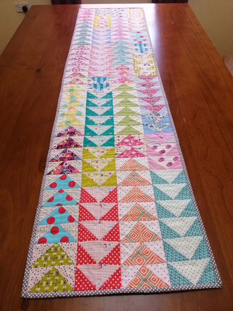 K&ouml;nnte beinhalten: Ein farbenfroher Patchwork-Tischl&auml;ufer mit einem geometrischen Muster aus Dreiecken. Der L&auml;ufer besteht aus verschiedenen Stoffen in den Farben Rosa, Gr&uuml;n, Blau, Gelb und Rot. Der L&auml;ufer ist etwa 46 cm breit und 183 cm lang.