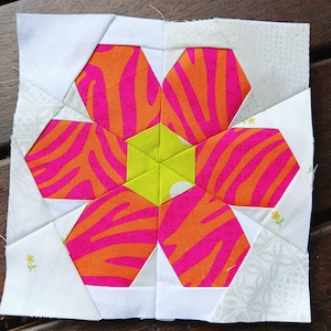 Op de afbeelding: Een wit vierkant van stof met een roze, oranje en gele bloemdessin. De bloem heeft zes bloemblaadjes en een geel centrum.