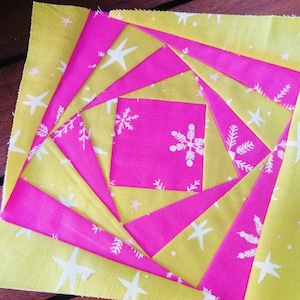 Op de afbeelding: Een roze en gele stoffen quiltblok met een spiraalvormig ontwerp. De stof heeft een wit sneeuwvlokkenpatroon op de roze stof en een wit sterrenpatroon op de gele stof.