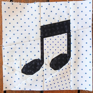 May include: A white fabric square with a blue polka dot pattern. A black musical note is sewn onto the fabric.