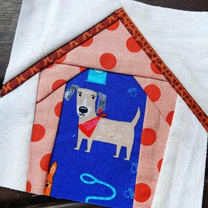 May include: A white fabric square with a brown and orange border, depicting a dog house with a red and white polka dot roof. Inside the house is a blue fabric with a brown dog and paw prints.