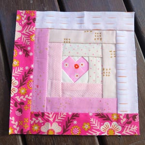 May include: A square fabric quilt block featuring a central pink heart surrounded by concentric strips of fabric in shades of pink, cream, and white. The outer border consists of a vibrant hot pink floral print with yellow and white flowers.