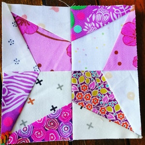 Könnte beinhalten: Ein Patchwork-Quiltblock mit einem Sternmuster. Der Block besteht aus verschiedenen Stoffen in Rosa-, Lila- und Weißtönen. Die Stoffe haben unterschiedliche Muster, darunter florale, geometrische und abstrakte Designs.