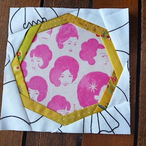 Op de afbeelding: Een wit vierkant van stof met een geel en wit gepatroneerd zeshoek in het midden. De zeshoek is omgeven door een gele rand. De zeshoek heeft een roze en wit stof met een herhalend patroon van vrouwengezichten met roze haar.