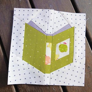 May include: A square quilt block featuring a central open book design made of solid olive green fabric. The pages are white with a small apple print. The background is white fabric with small grey polka dots, and a light purple accent strip.