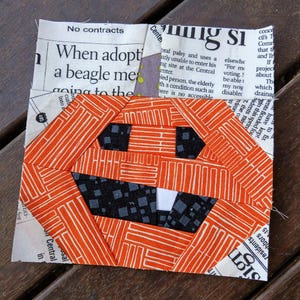 May include: A handmade orange and white fabric pumpkin with a black and grey mouth and eyes, and a single white tooth. The pumpkin is made of geometric shapes and is set against a background of newspaper clippings. The text "No contracts" is visible.