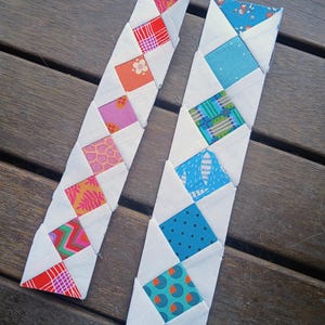 May include: Two handmade fabric bookmarks with colourful geometric patterns. One bookmark features red, orange, and pink squares, while the other has blue and green squares. Each square is set in a white background.