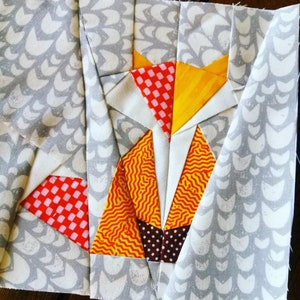 May include: A patchwork quilt block featuring a fox design. The fox is made of various fabrics in red, white, yellow, and brown. The background fabric is gray with a white pattern.