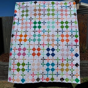 May include: A handmade quilt with a white background and a repeating pattern of colorful diamond shapes. The diamonds are in various colors, including pink, green, orange, and blue. The quilt is hanging outdoors.