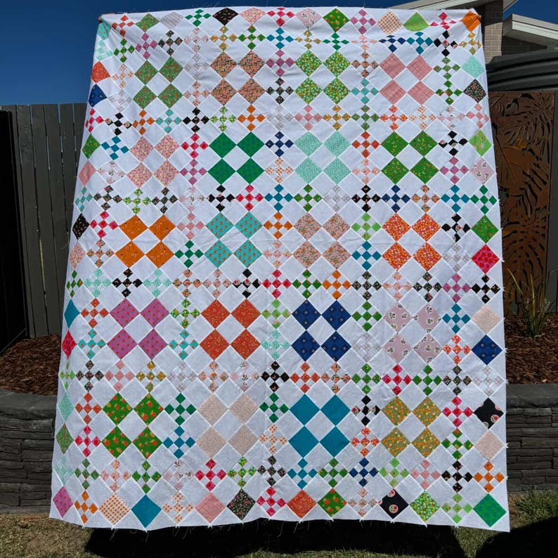 May include: A handmade quilt with a white background and a repeating pattern of colorful diamond shapes. The diamonds are in various colors, including pink, green, orange, and blue. The quilt is hanging outdoors.