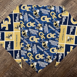 May include: A blue and gold pet bandana featuring the words "GEORGIA TECH" and the letters "GT". The bandana has a repeating pattern of the Georgia Tech Yellow Jacket mascot and logo. The bandana is made of fabric.