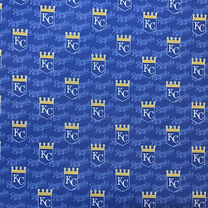 May include: Blue fabric with a repeating pattern of the Kansas City Royals logo. The logo features a gold crown above the letters "KC" inside a white shield. The word "Royals" is also printed on the fabric.