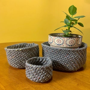 May include: Three gray crocheted plant pots, one inside the other, with a small potted plant in the largest pot. The plant has green leaves and is in a white ceramic pot with a colorful pattern.